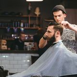 Young handsome barber making haircut of attractive bearded man in barb